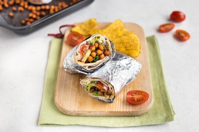  Una envoltura de ensalada de garbanzos picante enrollada en papel de aluminio y cortada por la mitad en una tabla de cortar.