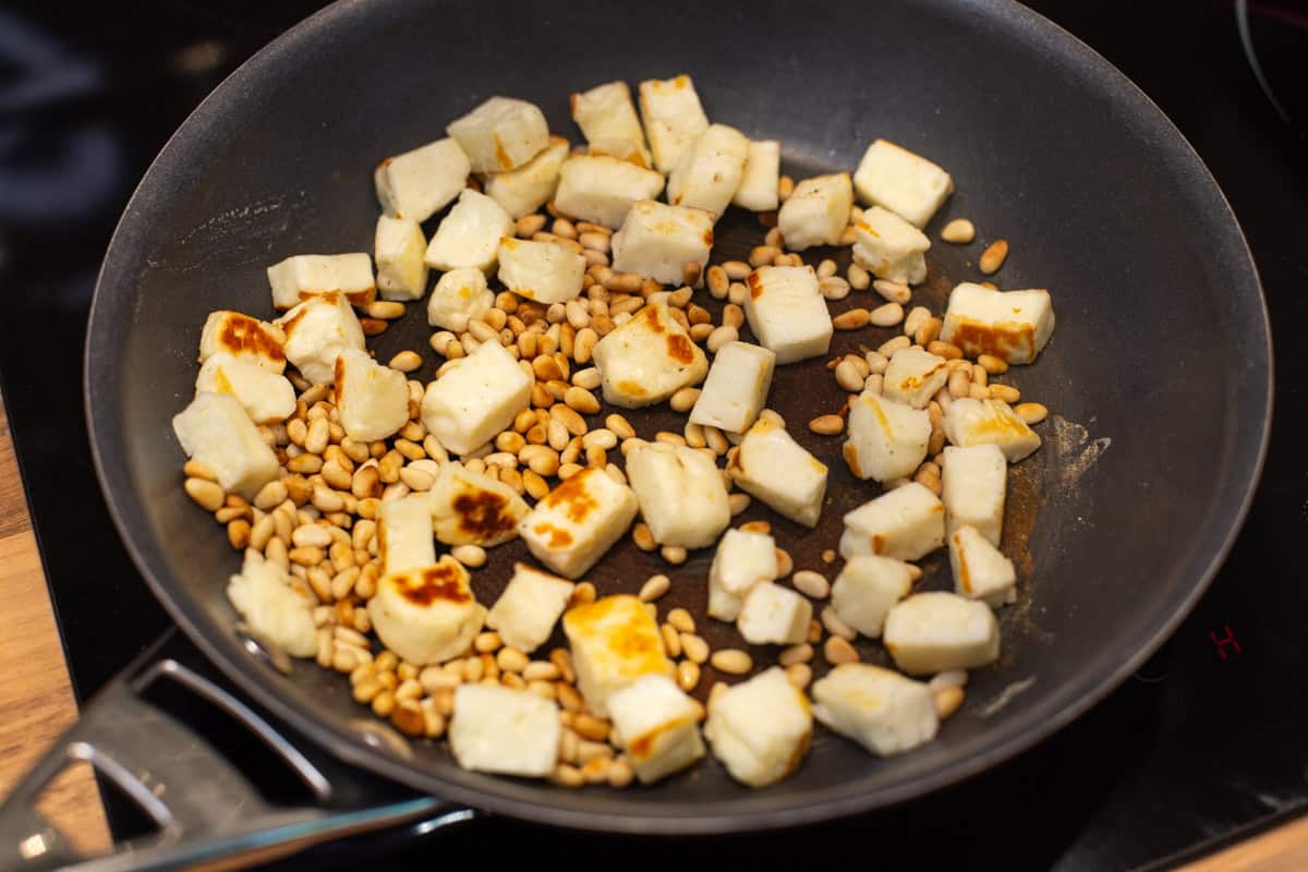 Diced halloumi and pine nuts toasting in a frying pan.