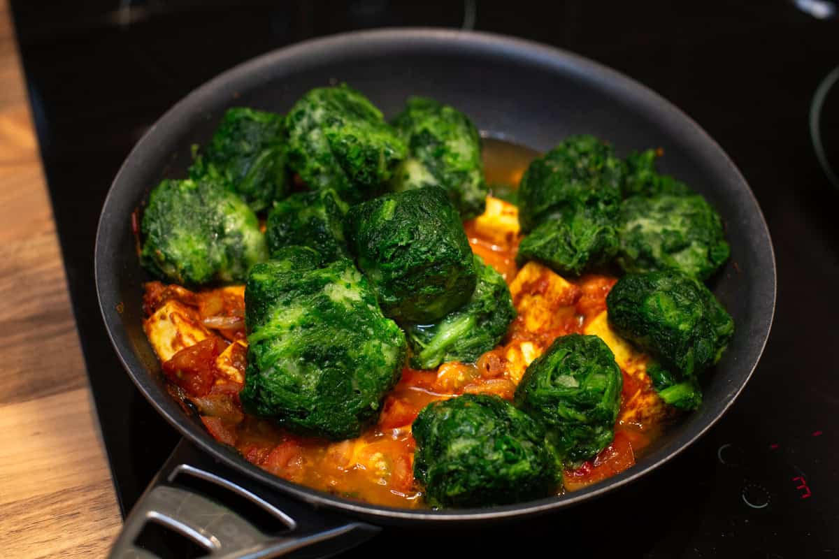 Blocks of frozen spinach added to a frying pan with a tomato mixture.