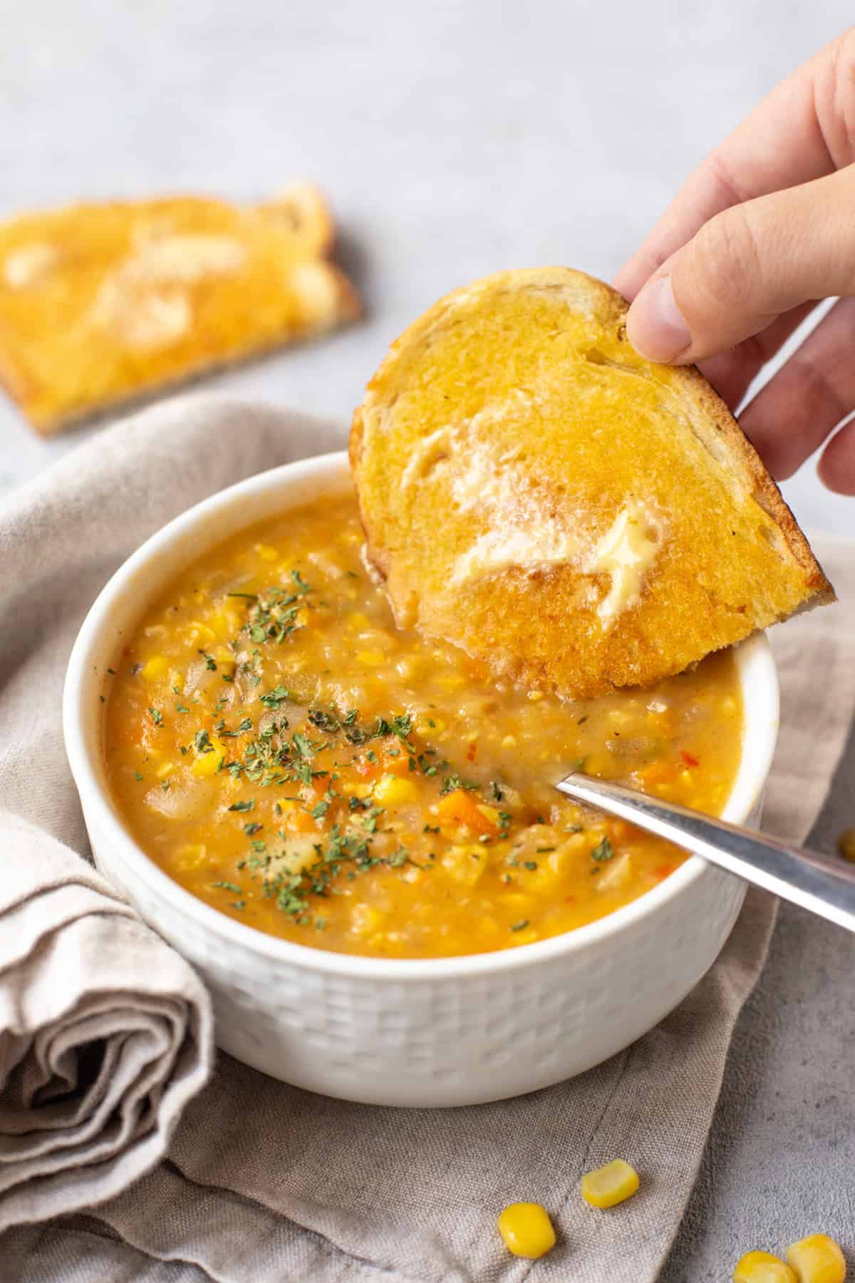 A piece of buttered toast being dipped into vegan corn chowder.