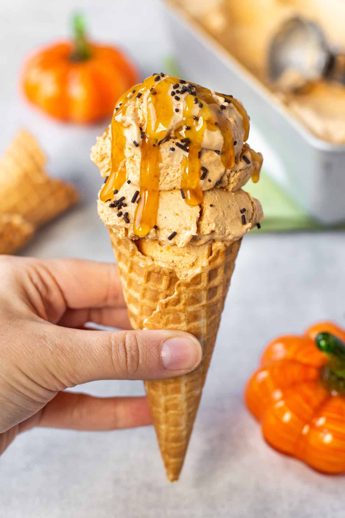 A hand holding a cone filled with pumpkin pie ice cream and topped with sprinkled and caramel sauce.