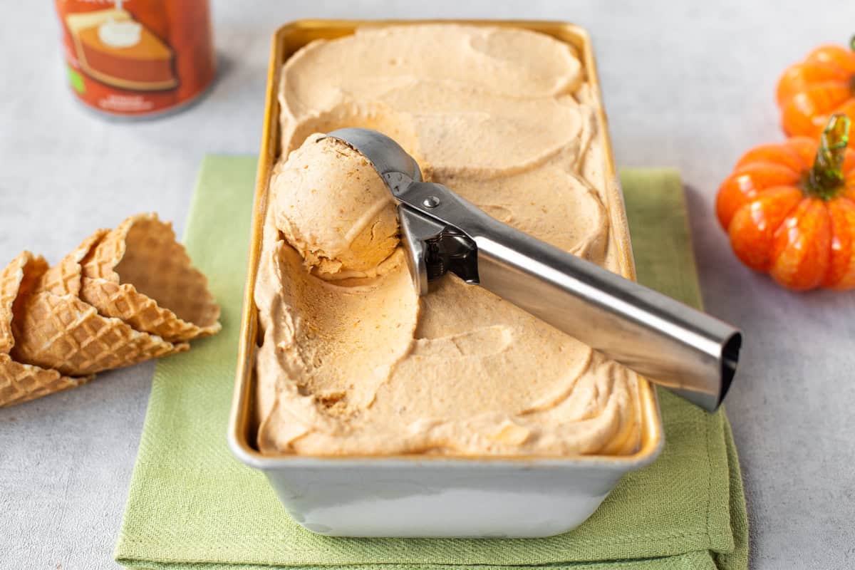 A metal ice cream scoop taking a spoonful of pumpkin pie ice cream from a loaf tin.