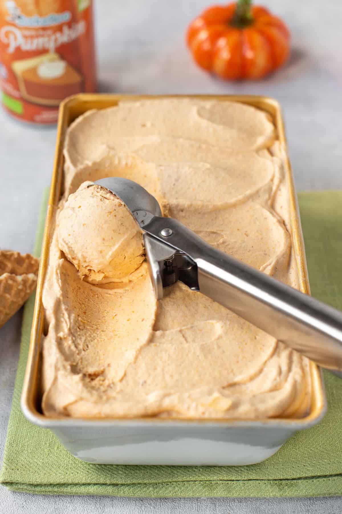 A metal ice cream scoop taking a spoonful of pumpkin pie ice cream from a loaf tin.