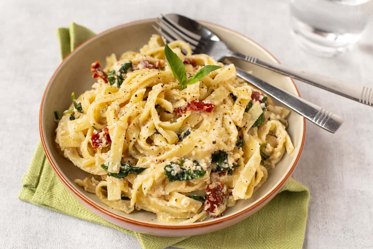 A bowl of creamy tofu pasta with spinach and sun-dried tomatoes.